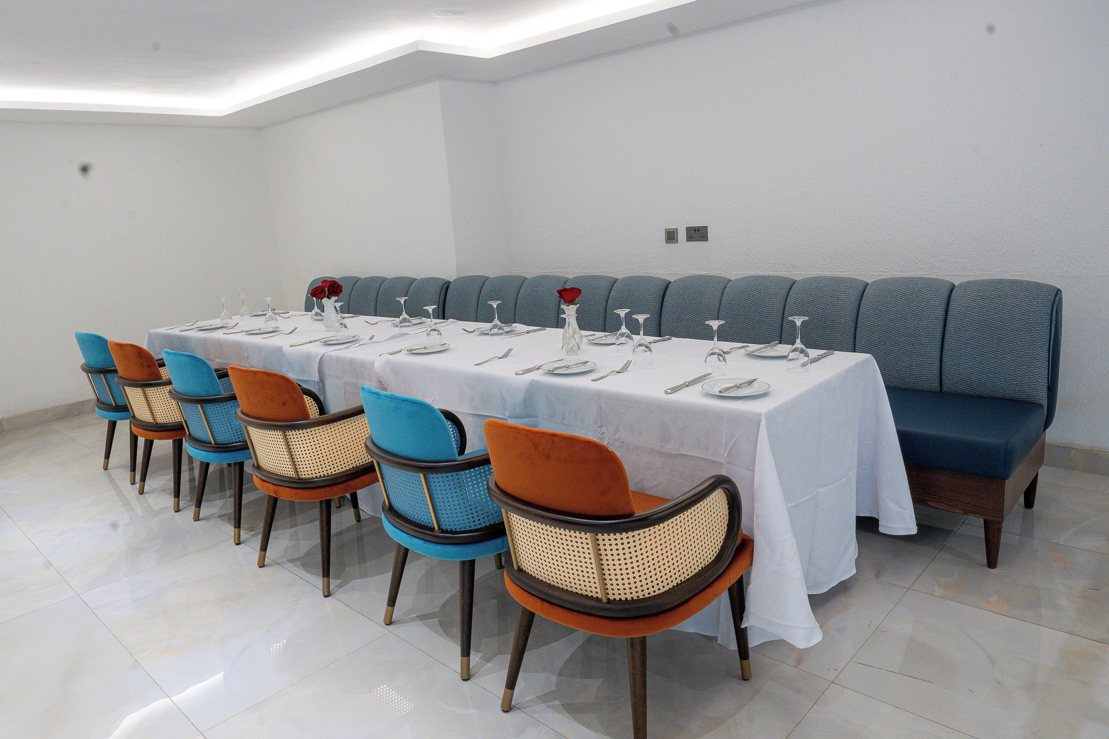 Uma sala de jantar com uma mesa comprida posta para refeição, rodeada por cadeiras com almofadas azuis e laranja, e um banco acolchoado em azul.
