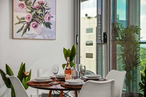 A dining table set with plates, glasses, and a bottle of wine, with a large window and a painting of flowers in the background. - 2BR Urban Jungle Escape • Walk to Everything (Melbourne)