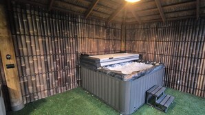 A hot tub with a wooden bench, surrounded by bamboo walls and artificial grass. - Family Luxury Country Retreat Pool & Spa – Lodge 2 (Yarrambat)