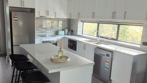 A modern kitchen with white cabinets, a large island with bar stools, a stainless steel refrigerator, and a dishwasher. - Family Luxury Country Retreat Pool & Spa – Lodge 2 (Yarrambat)