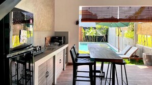 A modern kitchen with a bar area, a dining table with chairs, and a view of a swimming pool and garden. - Villa Loba (Denpasar)