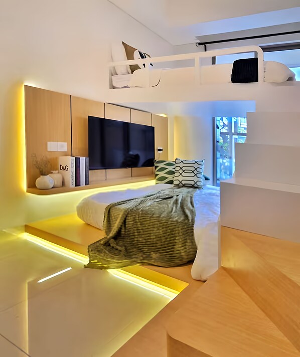 Une chambre moderne avec un lit superposé, un téléviseur à écran plat, une table de chevet et une étagère intégrée où sont rangés des livres et des objets décoratifs.