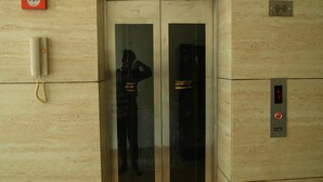Egy üvegajtós lift, egy piros színű vészhelyzeti telefon és egy piros gombbal ellátott kezelőpanel.