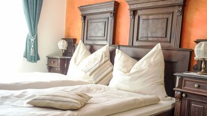 Una cama bien tendida con sábanas blancas y almohadones decorativos, una mesita de noche de madera con una lámpara, y un cabecero de madera oscura sobre una pared anaranjada.