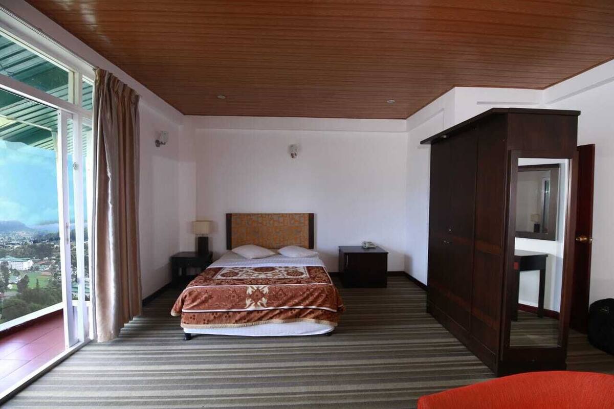A hotel room with a large bed, a wardrobe, a nightstand, a lamp, and a view of the city.