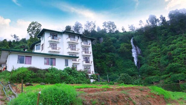 A white multi-story building with balconies, surrounded by lush greenery and a waterfall. - Ashley Resorts Nuwaraeliya (Nuwara Eliya)