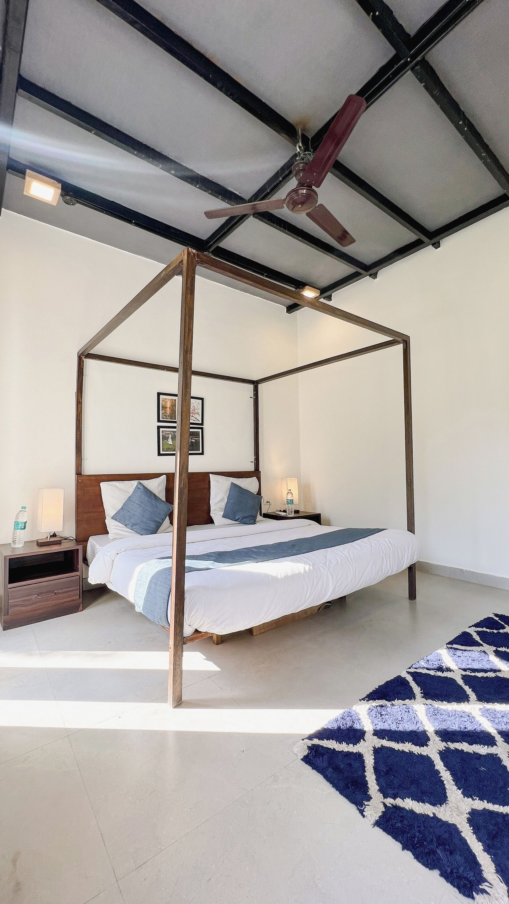 A four-poster bed with a wooden frame, white bedding, and two pillows. There is a nightstand with a lamp and a bottle of water. The room has a tiled floor and a blue patterned rug.