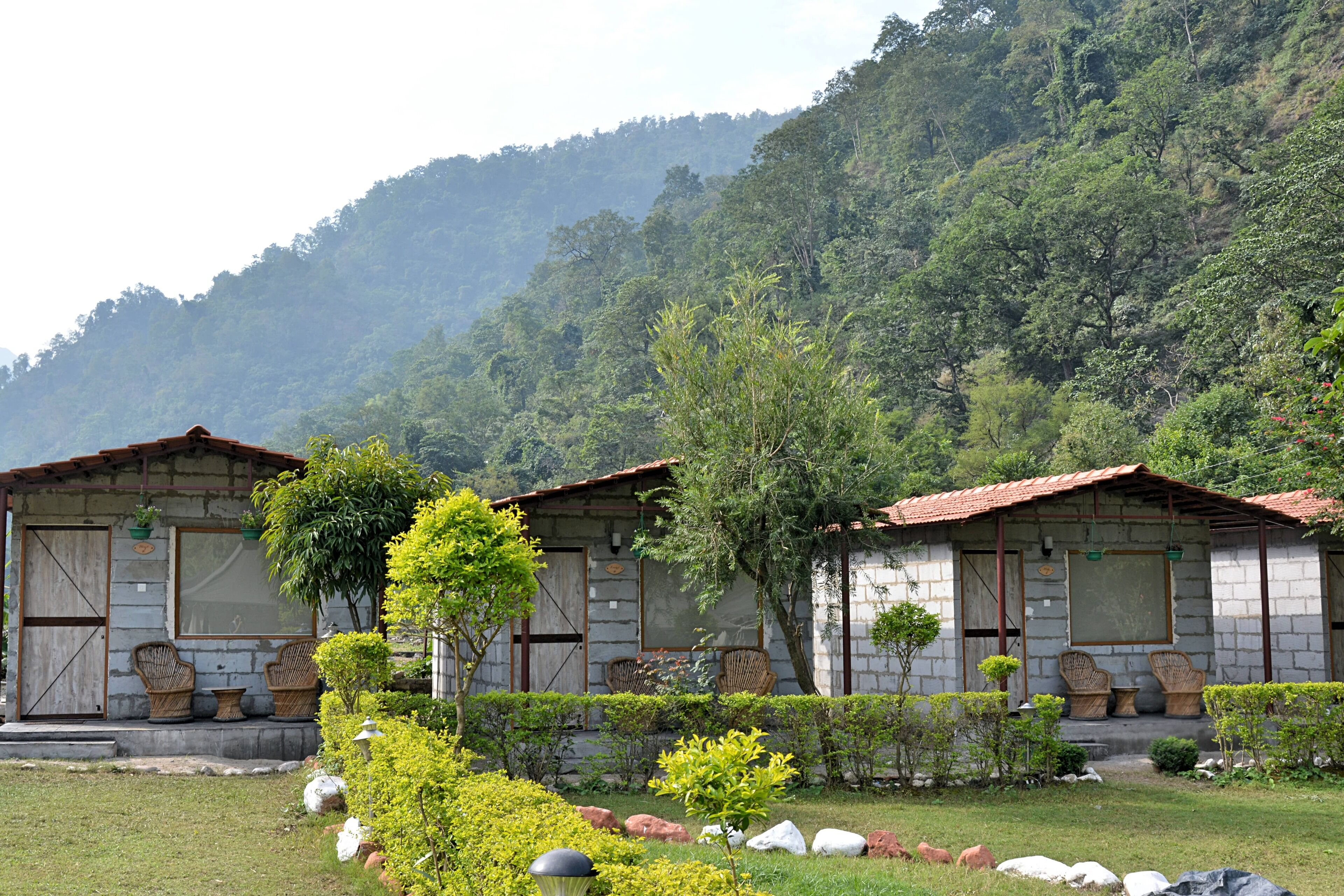 A rustic building with a stone facade, two wicker chairs on the porch, and a lush garden with various plants and rocks.