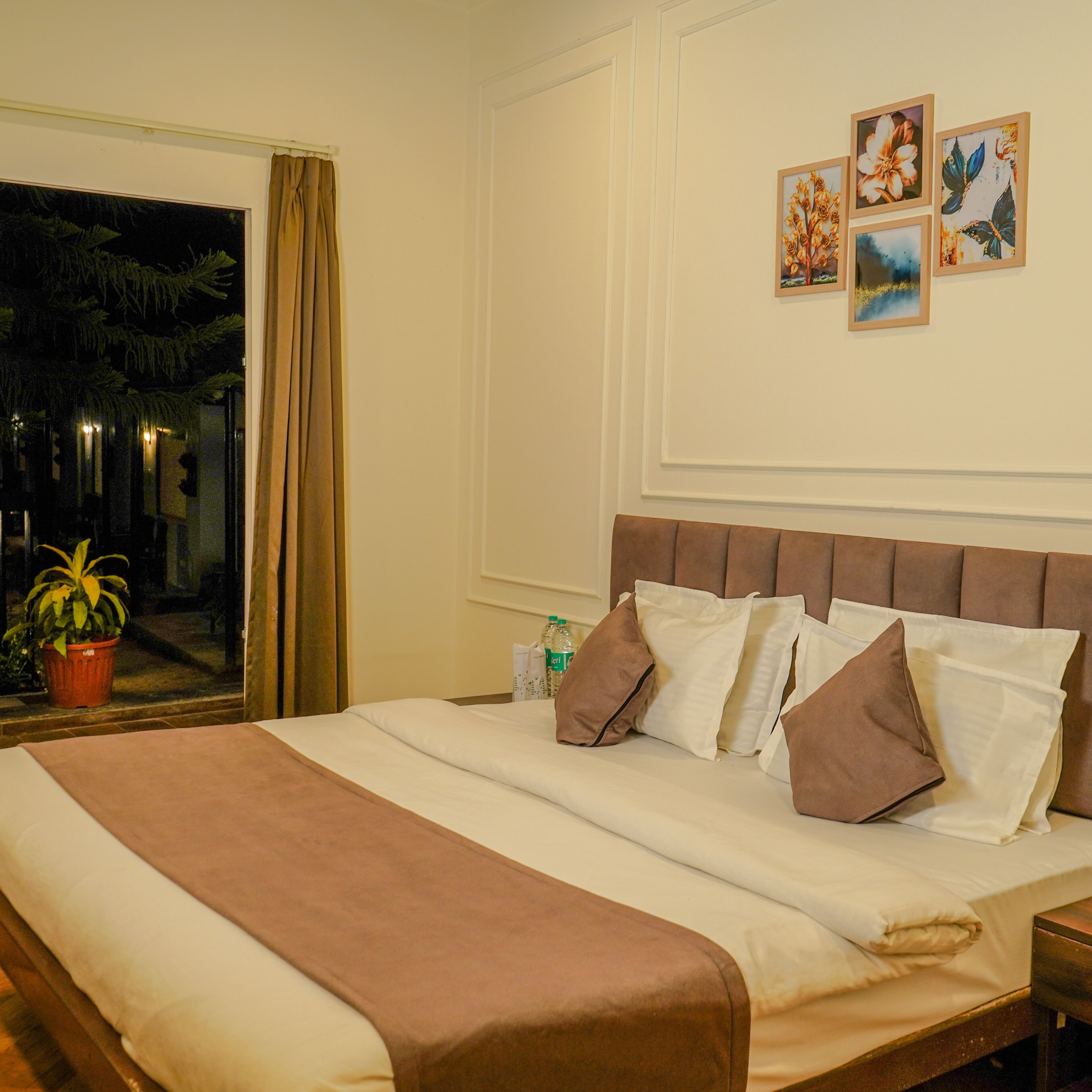 A hotel room with a large bed, a window with curtains, a potted plant, and wall art.