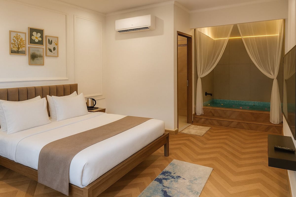 A hotel room with a bed, bedside table, air conditioning unit, artwork on the wall, and a bathroom with a shower and curtain.
