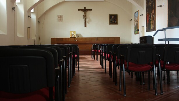 Interior de iglesia con hileras de sillas, altar de madera y una cruz fijada en la pared.