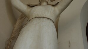 Una estatua blanca de gran tamaño con los brazos extendidos, frente a una pared que tiene un adorno de madera encima.