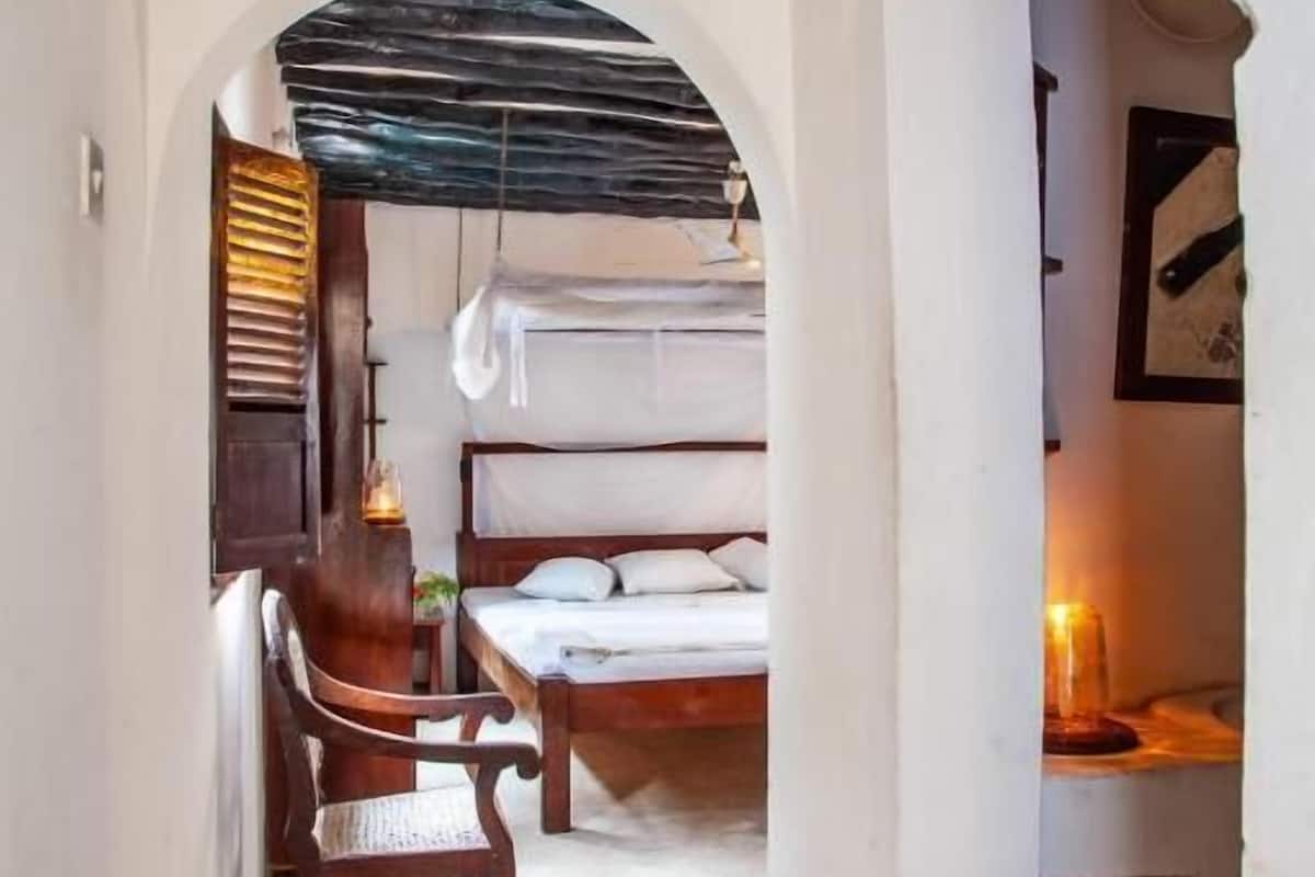 A narrow hallway with a wooden chair, a bed with a canopy, and a desk with a candle.