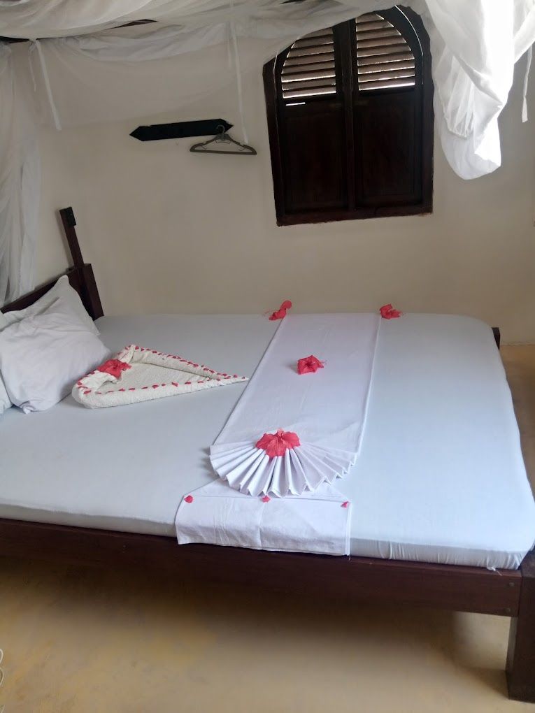 Ein ordentlich bezogenes Bett mit weißen Bettwäsche und rosa dekorativen Elementen, ein hölzerner Kopfteil, ein Fenster mit Jalousien und ein Wandhaken.