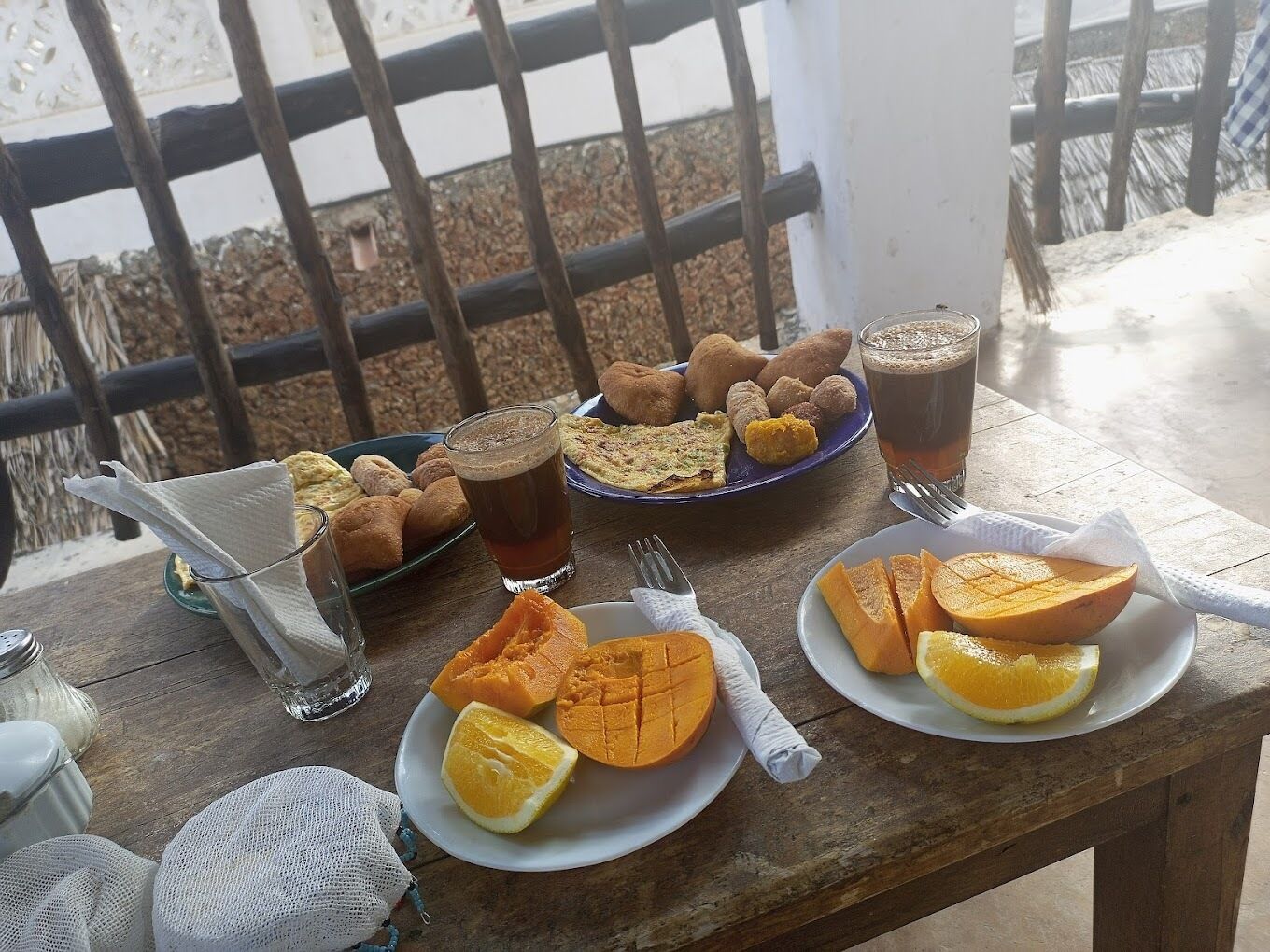 A wooden table with plates of food, including sliced fruits and pastries, and glasses of a dark beverage.