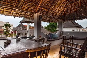 An outdoor seating area with a thatched roof, wooden furniture, and a view of surrounding buildings.