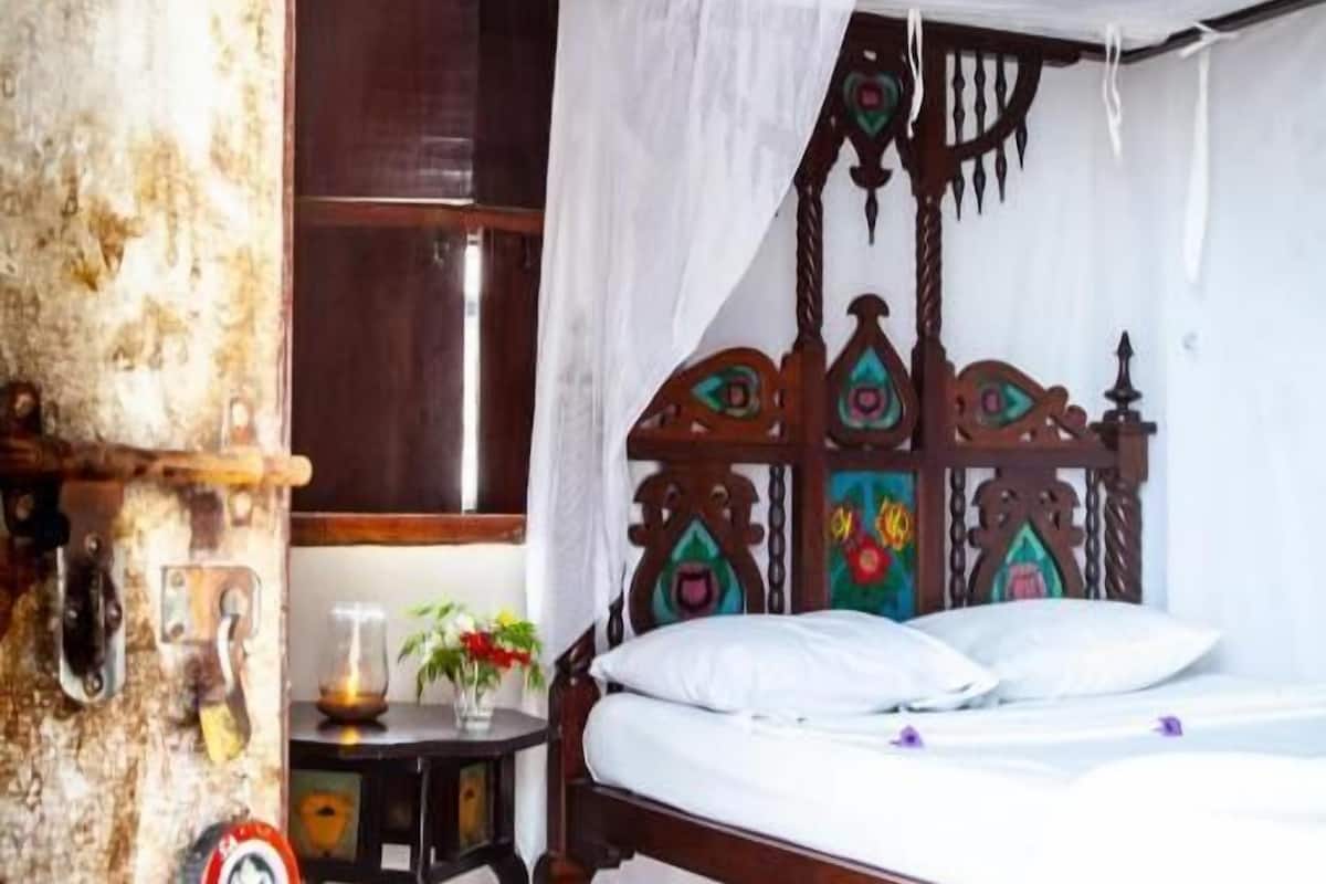 A room with a bed under a canopy, a wooden headboard, a small table with a vase, and a rustic wall.