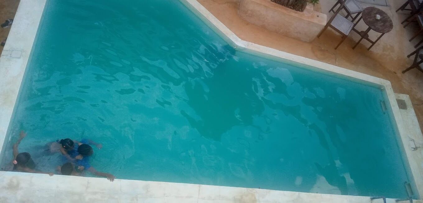 A swimming pool with clear blue water, surrounded by a tiled deck and a few outdoor tables and chairs.