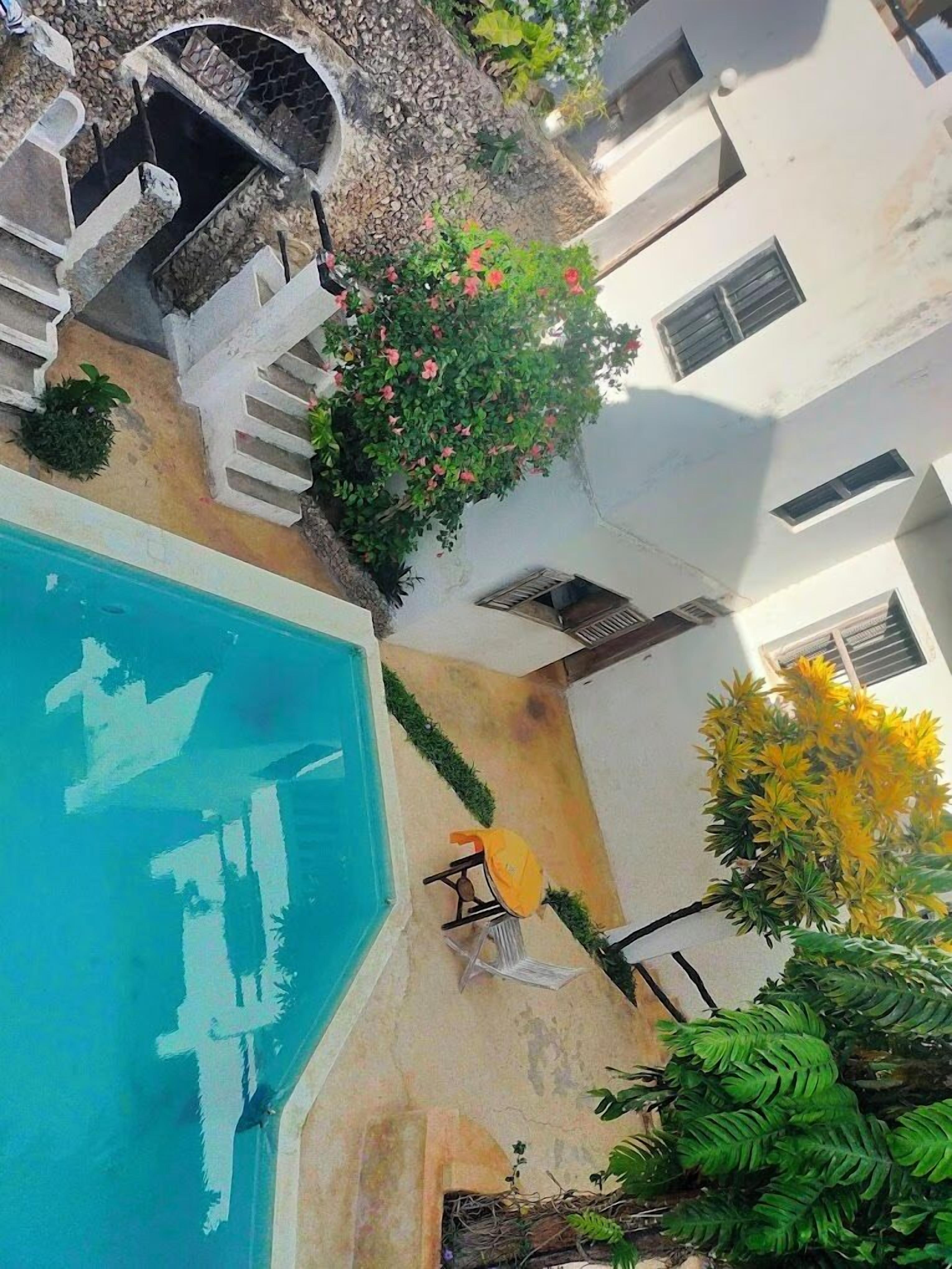 A swimming pool with a stone border, a small table with a chair, and a staircase leading to a white building.
