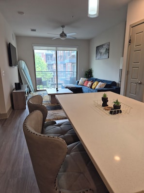 A modern living room with a dining area, a ceiling fan, a large window with a view of buildings, and a door. - Luxury Frisco with 2 queen beds sleeps 4 Universal kids resort, FIFA Stay! (Frisco)