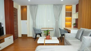 A modern hotel room with a wooden floor, a white sofa, a small table with a potted plant, a TV, and a desk with a chair. - Batuwa Homes (Kathmandu)