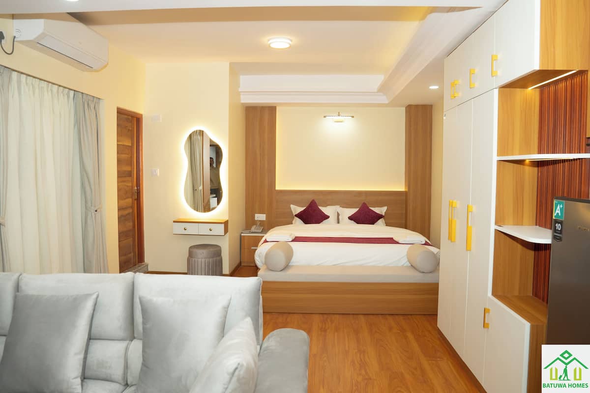 A modern hotel room with a bed, a sofa, a mirror, a wardrobe, and a built-in fridge.