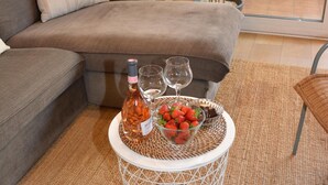 Un moderno salón con un sofá gris, una mesa redonda de centro con una botella y vasos, un tazón de fresas y una planta en maceta.