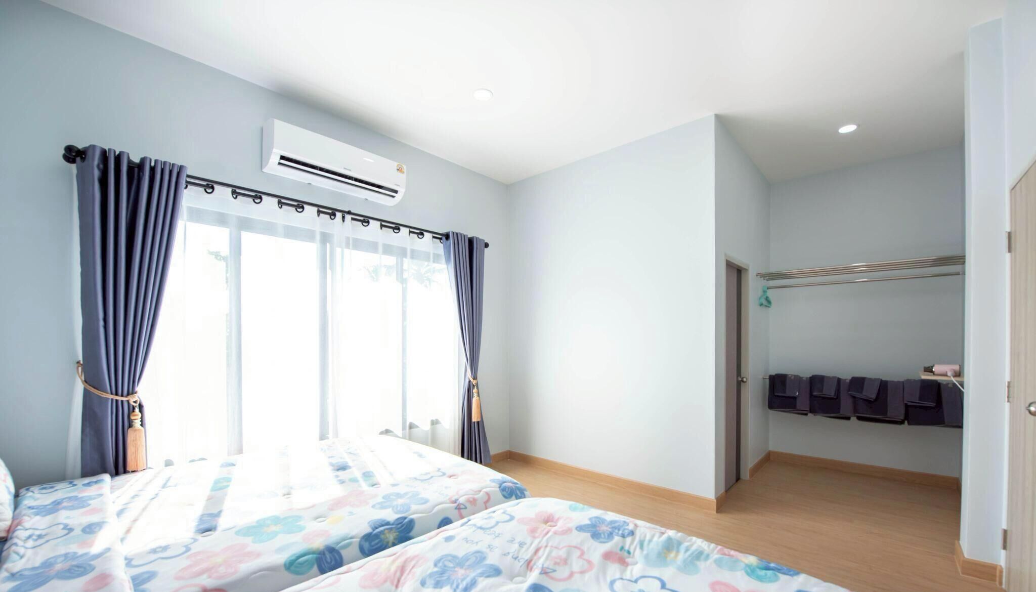 A hotel room with two beds, an air conditioning unit, a window with curtains, a door leading to a bathroom, and a rack with towels.