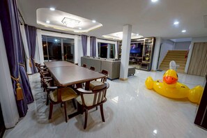 A spacious dining area with a long table and chairs, a large window with curtains, and a yellow inflatable duck.