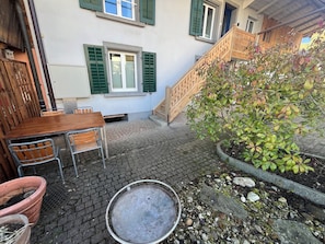 A courtyard with a wooden table and chairs, a metal chair, a potted plant, and a metal trash bin. - Boutique apartment in the Wynental valley in the triangle between Bern, Zurich and Lucerne. (Reinach)