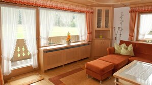 En hyggelig stue med en sofa, et glasbord og en radiator. Der er gardiner ved vinduerne, og et lille skab er synligt.