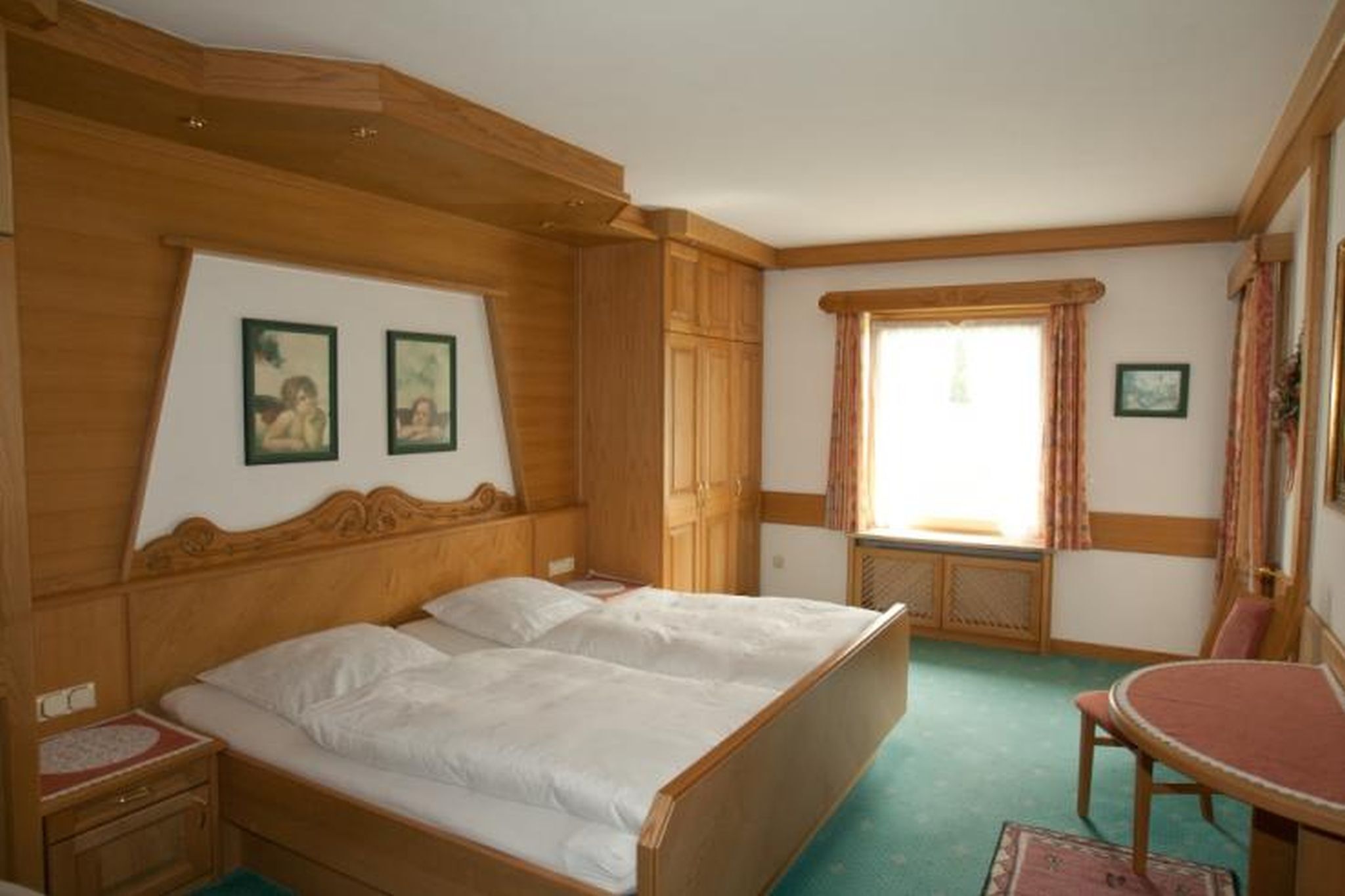 Ein Zimmer mit einem hölzernen Bett, einem kleinen Tisch und einem Fenster mit Vorhängen.
