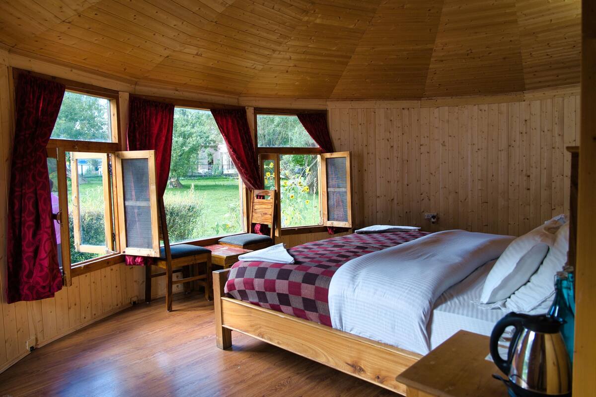 A wooden cabin bedroom with a bed, a desk, and two windows with curtains.