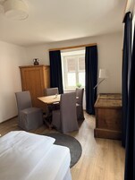 Una habitación de hotel con una mesa de comedor de madera, dos sillas grises, un armario de madera, una cama, una mesita de noche con una lámpara y una ventana con cortinas.
