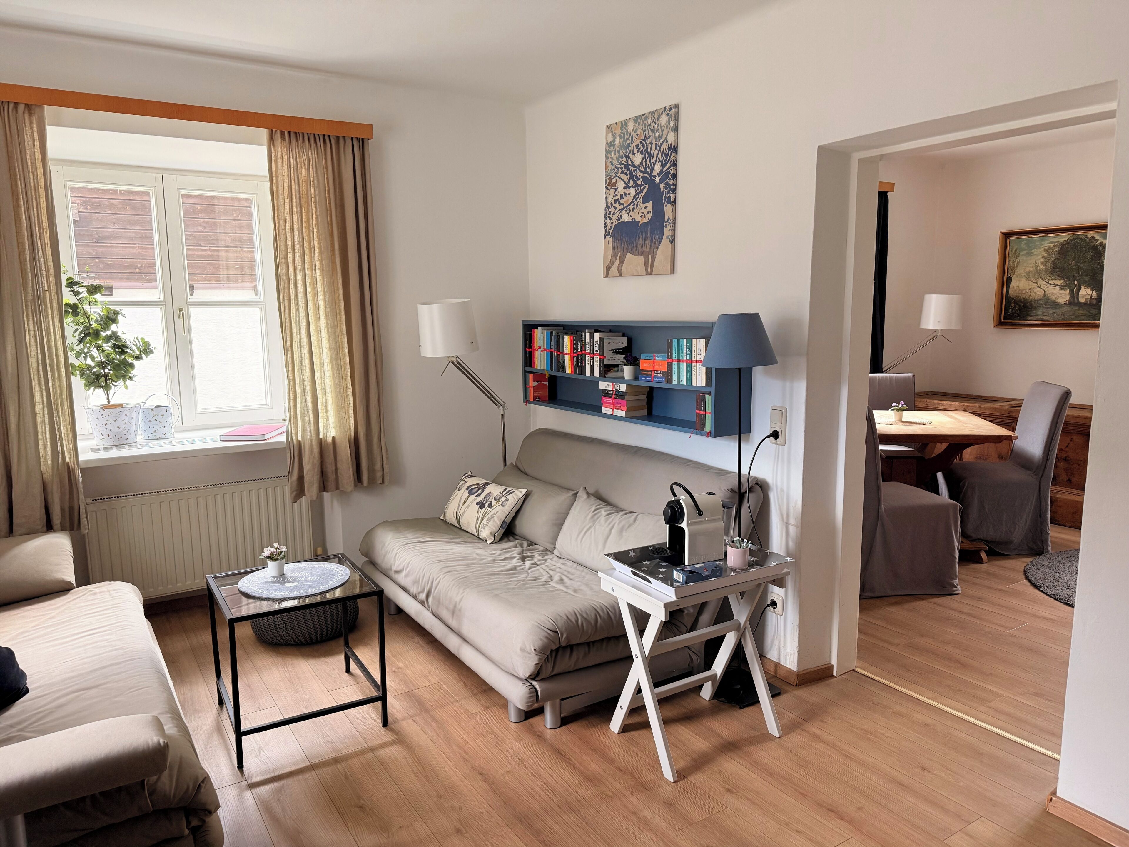 A living room with a sofa, a coffee table, a side table, a bookshelf, a desk with a lamp, a dining area with a table and chairs, and a window with curtains.