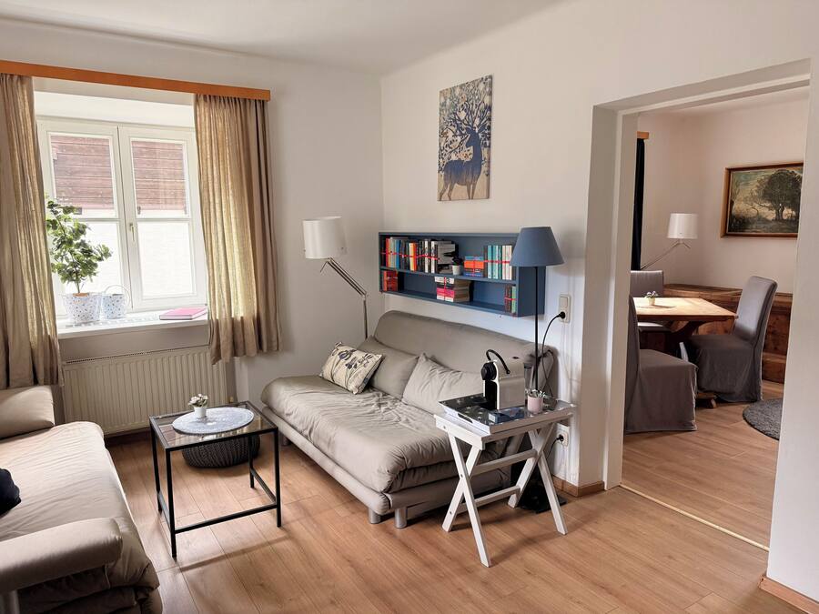 Una sala de estar con sofá, una mesa de centro, una mesita auxiliar, una estantería, un escritorio con lámpara, un área de comedor con mesa y sillas, y una ventana con cortinas.