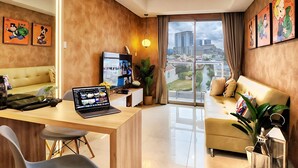 Un salon moderne avec un canapé, un bureau, un ordinateur portable et une télévision. Des tableaux encadrés ornent le mur, et la fenêtre offre une vue sur la ville.