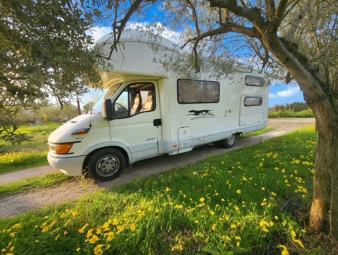 A white RV parked on a dirt path with yellow flowers and trees in the background.