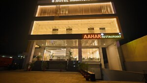 Un hôtel dont l’enseigne, bien visible, porte les noms « Hôtel Madhuram » et « Restaurant Aahar », et qui est particulièrement visible la nuit.