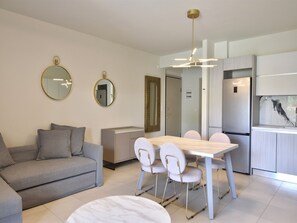 A modern living room with a sofa, a dining table with chairs, a kitchen area with a refrigerator, and two wall-mounted mirrors.