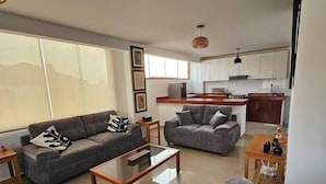 A modern living room with grey sofas, a glass coffee table, and a kitchen area with white cabinets and a wooden countertop.