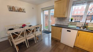 A modern kitchen with wooden cabinets, a dishwasher, and a dining area with a glass table and chairs. - Charming 5-Bedroom Townhouse in Central Gloucester (Gloucester)