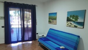 Ein Zimmer mit einem blau gestreiften Sofa, einem Fliesenboden, einer vor einer Gardine liegenden Tür und zwei Wandbildern.