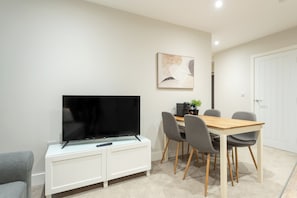 A modern living room with a flat-screen TV on a white cabinet, a dining table with four chairs, and a painting on the wall.