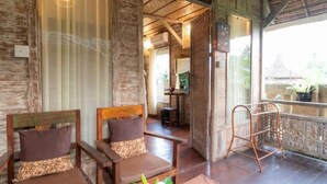 A wooden room with two cushioned chairs and a small table, a balcony with a metal railing, and a view of the outside.