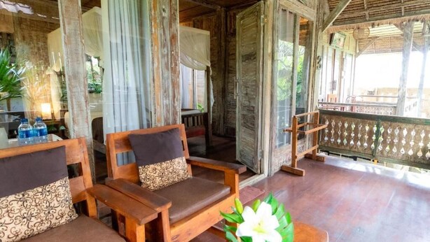 A wooden porch with two wooden chairs and a table, a railing, and a view of the outside.