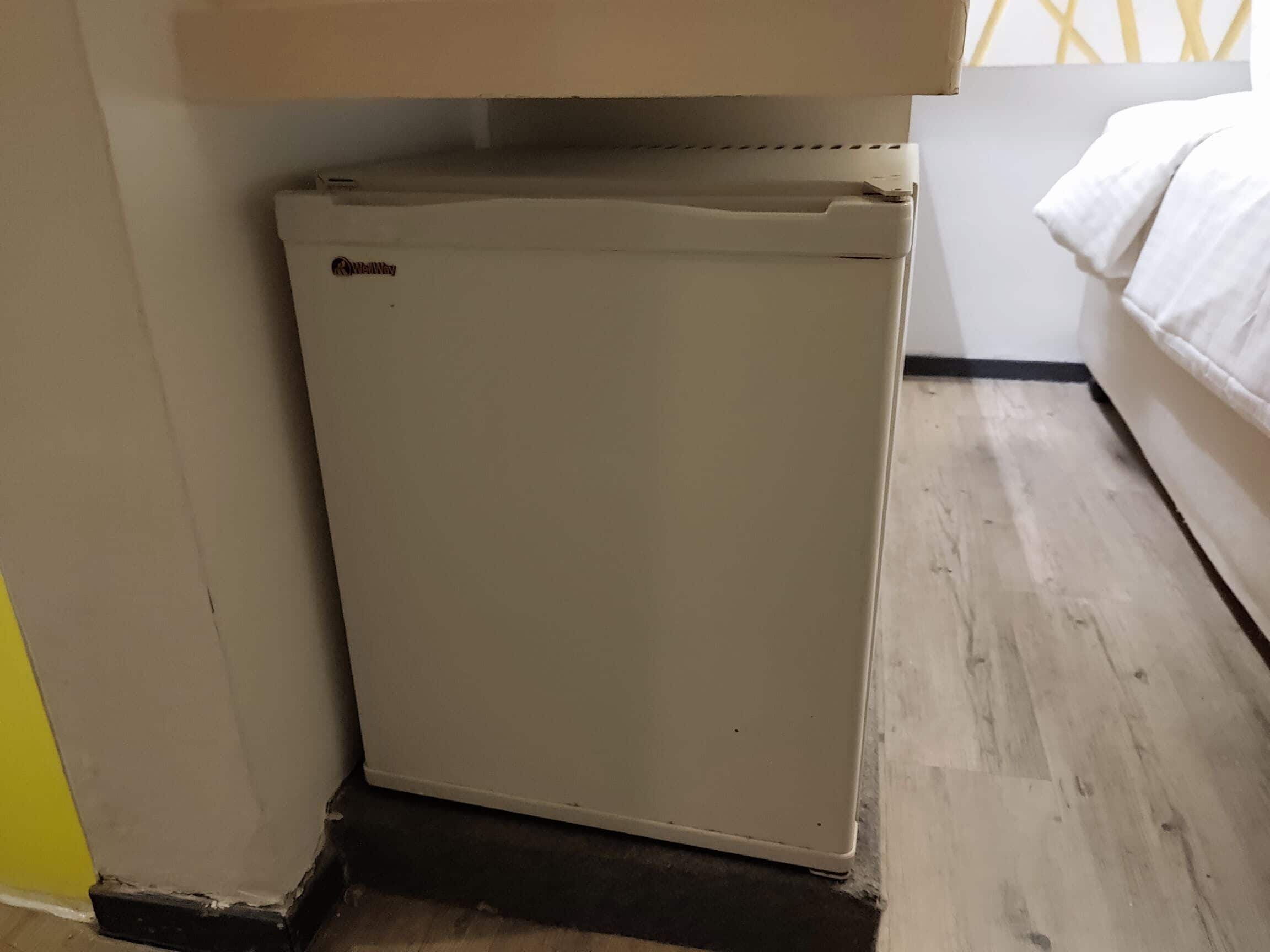 A compact refrigerator in a room with a wooden floor and a bed with white bedding.