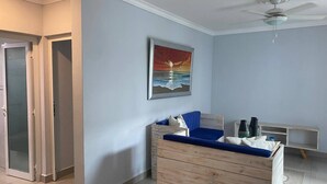 Een kamer met een houten dressoir, een blauwe bank, een schilderij van een zonsondergang boven de oceaan en een plafondventilator.