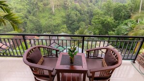 A balcony with two wooden chairs and a table, overlooking a lush green forest. - Deluxe room upper floor with pool & nature view (Bali)