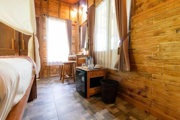 A wooden cabin room with a bed, a desk, a chair, and a trash bin.